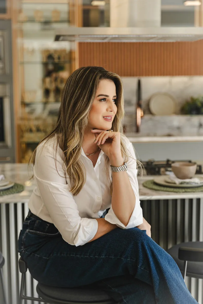 Mariana Carmo sentada em uma banqueta na cozinha, mão no queixo, com camisa branca