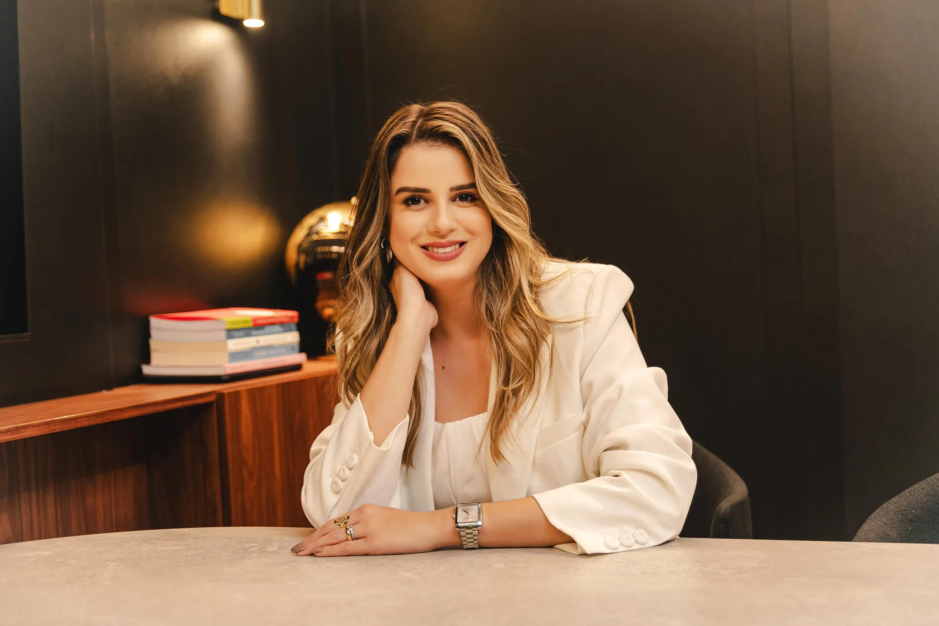 Mariana Carmo, psicóloga especialista em compulsão alimentar e comer emocional, sorrindo, sentada à mesa com blazer branco